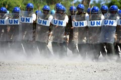 A row of UN Police officers wearing blue helmets and holding shields with the letters UN on the front.