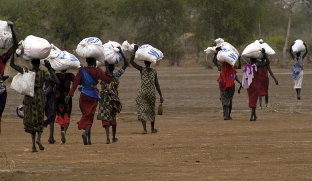 A line of women walk across dusty ground carrying large bags