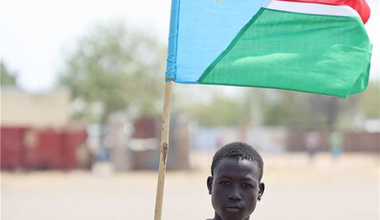 A boy standing in front of the South Sudanese flag. 