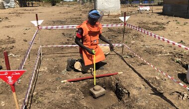 A person wearing demining personal protective equipment in a patch of land sealed off by red tape and warning signs removes an unexploded ordnance from the ground.