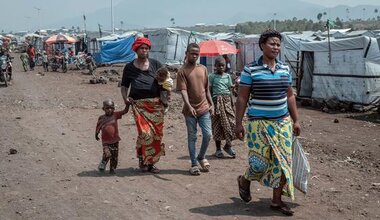 A group of people walk along a dirt round, through a camp for displaced people 